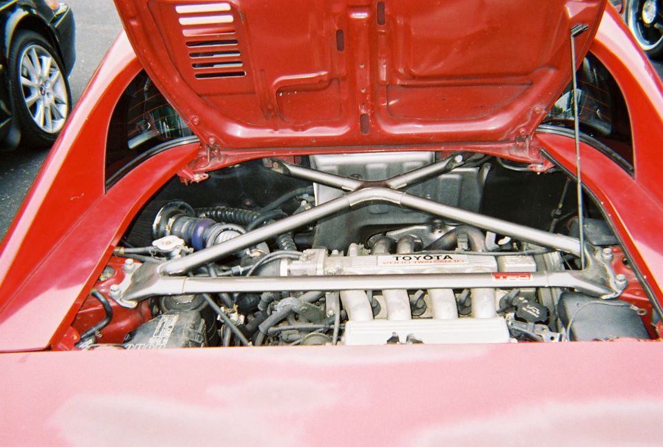 Red Toyota MR2 engine bay with cross brace and Toyota engine cover. Another car is partially visible on the left