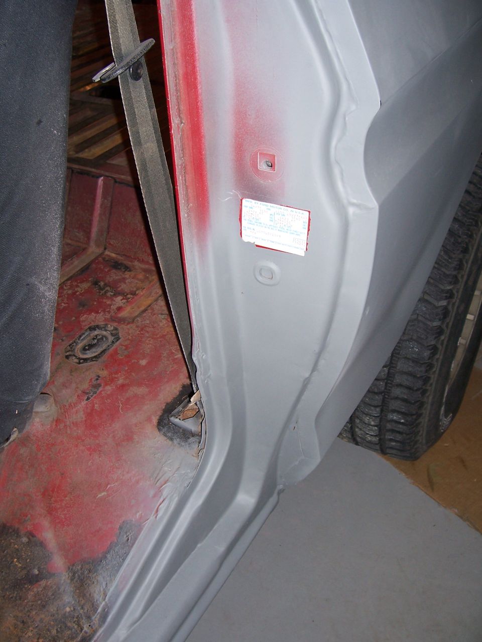 Interior of a vehicle, showing the door jamb area with a seatbelt and a sticker on the door frame. The floor is red and appears rusty