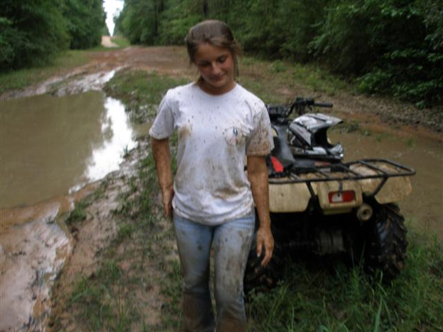 Mud-covered person standing beside a yellow ATV on a flooded dirt trail