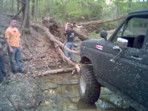 1986 Ford Bronco - A person uses an axe to clear a fallen tree blocking a muddy path where a Ford Bronco is driving.