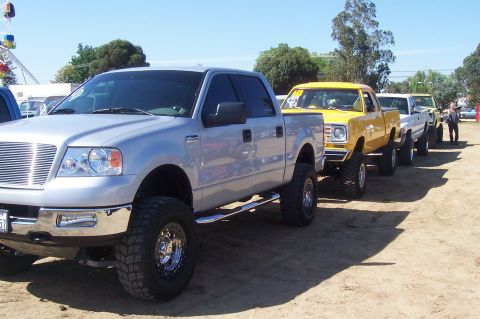 1994 Ford F-150 - A line of modified pickup trucks, including a silver Ford and a yellow Dodge, are parked outdoors on a sunny day.