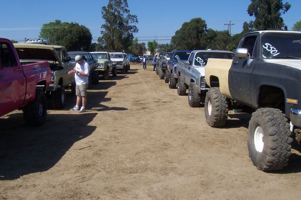 Several lifted pickup trucks parked in a dirt lot, some with hoods open. One truck is black and tan with huge tires
