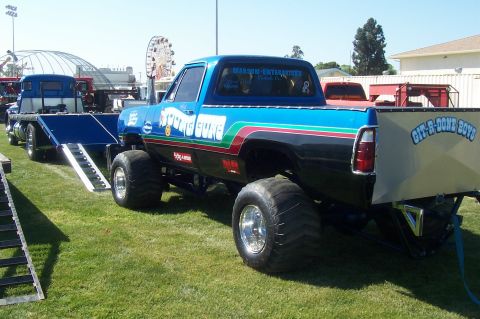 1994 Ford F-150 - This heavily modified truck, possibly a Dodge, is equipped with massive tires and custom graphics, and is shown towing a flatbed trailer.