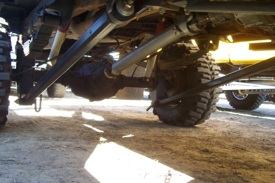 Undercarriage view of a lifted truck showing suspension and drivetrain components. Another vehicle is visible in the background