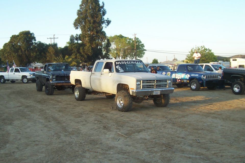 Several lifted pickup trucks parked in a dirt lot, one white crew cab in foreground