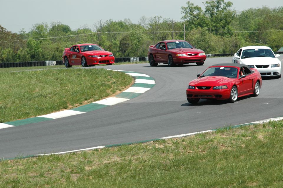 Four Ford Mustangs on a race track, three red and one white. The closest car is a red convertible