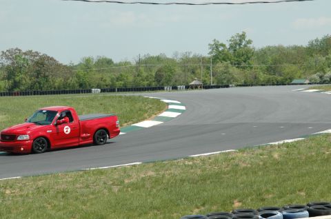 1993 Ford Mustang - A red pickup truck drives around a corner on a race track with green grass and trees in the background.