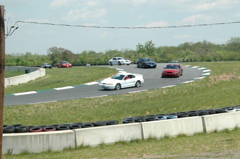 Several cars, including a white Ford Mustang, race on a track. A red car is in the foreground