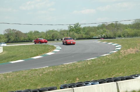 1993 Ford Mustang - A red 1993 Ford Mustang navigates a turn on a sunny day at the racetrack, with other vehicles visible in the background.
