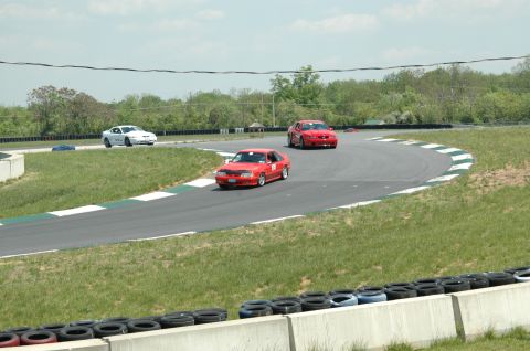 1993 Ford Mustang - Three cars are racing on a paved track with grassy shoulders and tire barriers.