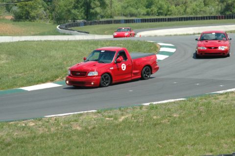 1993 Ford Mustang - A red pickup truck navigates a turn on a race track, with two other red vehicles visible in the distance.