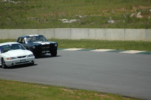 1993 Ford Mustang - Two Ford Mustangs, one modern and one classic, are on a paved race track with grassy banks and a concrete barrier.