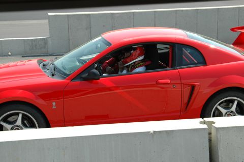 1993 Ford Mustang - A red coupe with a racing driver is seen from the side on a track. The vehicle's body style suggests it is a 2000 Ford Mustang.