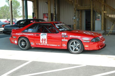1993 Ford Mustang - A red 1993 Ford Mustang Cobra race car, number 99, is parked under a covered area with other vehicles present.