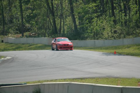 1993 Ford Mustang - Red Ford Mustang driving on a race track with trees and concrete barriers in the background.