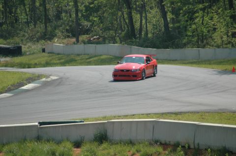 1993 Ford Mustang - A red sports car navigates a turn on a paved race track surrounded by trees.