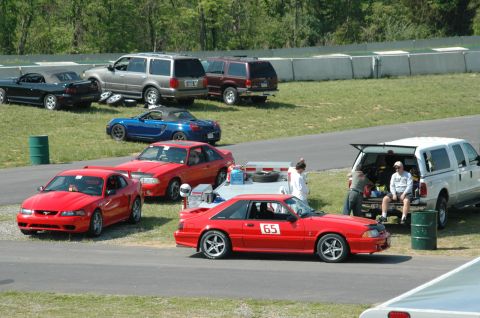 1993 Ford Mustang - A red coupe with racing number 65 is parked on the track, with several other vehicles and people in the background.
