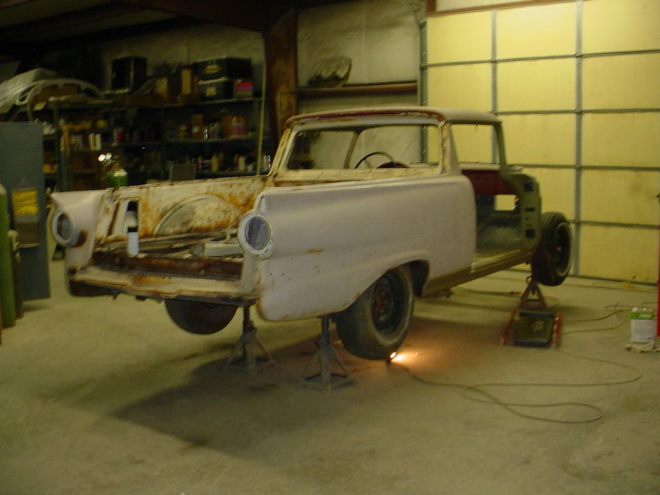 Rusty white Ranchero body shell on jack stands mid-restoration in a garage.