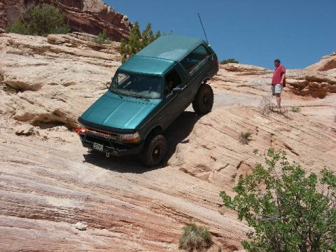 1996 Ford Bronco - A teal SUV navigates a steep, rocky trail with a person observing from above.