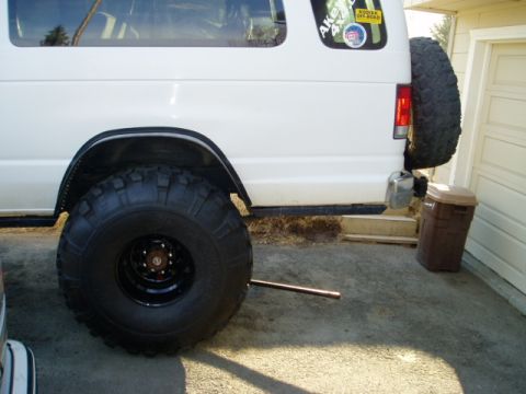 2000 Ford E350 - Side view of the rear corner of a white lifted van showing an oversized mud-terrain tire on a black steel wheel tucked under a flared fende…