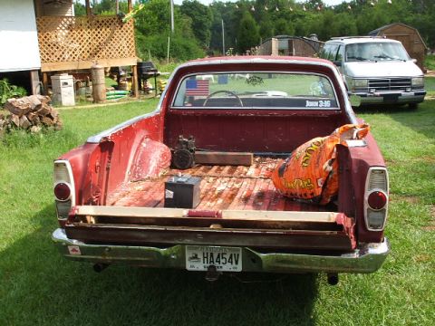 1966 Ford Ranchero - Rear view of a faded red Ford Ranchero parked on grass in a backyard, its bed floor surface-rusted and holding a battery, a small motor and…