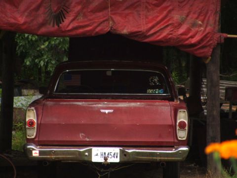 1966 Ford Ranchero - Rear view of a faded maroon Ford Ranchero coupe-utility parked under a red tarp shelter, with an American-flag decal in the rear window and…