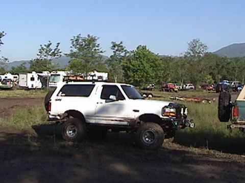 1996 Ford Bronco - A white SUV is parked on a dirt path with trees and RVs in the background.
