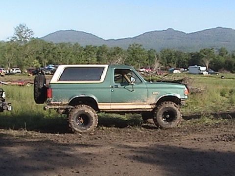 1996 Ford Bronco - A green Ford Bronco with a white top is parked in a muddy field with trees and hills in the background.