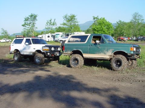 1996 Ford Bronco - Two modified Ford Broncos are parked side-by-side on a muddy off-road course, with RVs and trees visible in the distance.
