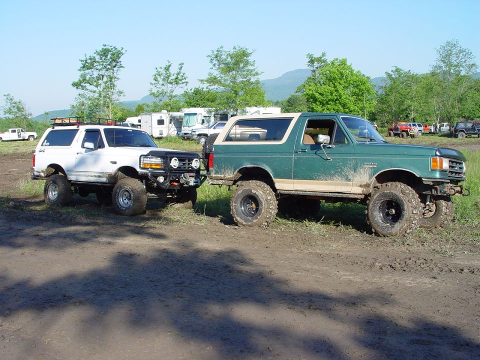 Two lifted Ford Broncos, one white and one green, parked on a dirt track with RVs and trees in the background