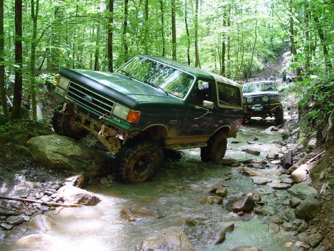 1996 Ford Bronco - Two off-road vehicles traverse a water-filled, rocky forest trail.