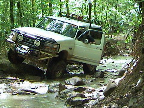1996 Ford Bronco - This white SUV navigates a challenging off-road path through a shallow, rocky stream surrounded by trees.