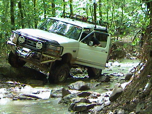 White SUV driving through a rocky stream in a forest. Driver's door is open