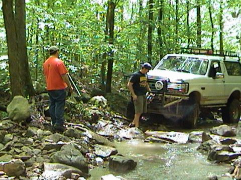 1996 Ford Bronco - A white Ford Bronco navigates a rocky stream in a wooded area, with two people nearby.