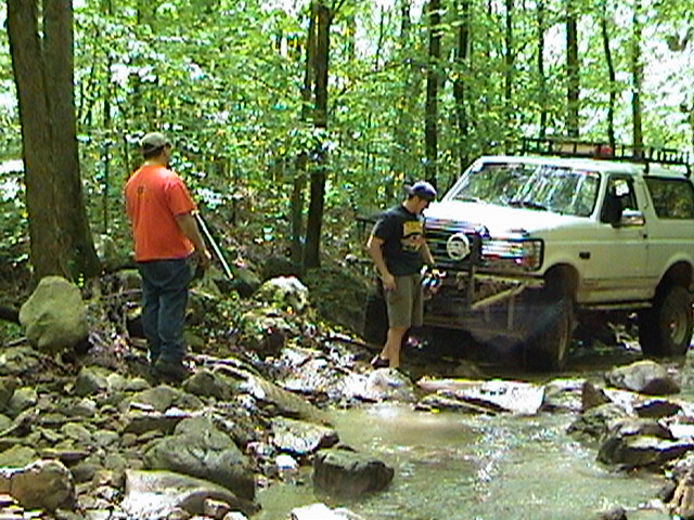 White Ford Bronco with roof rack driving through a rocky stream in a forest