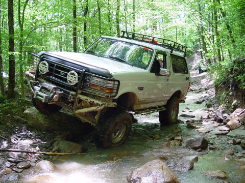 1996 Ford Bronco - White 1996 Ford Bronco off-roading through a shallow stream in a forest.