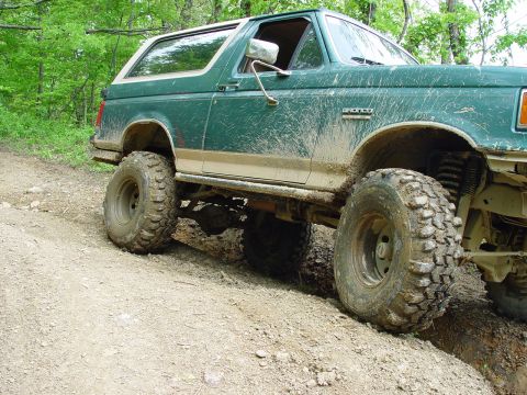 1996 Ford Bronco - Mud-covered green Ford Bronco navigates a rough dirt trail with lifted suspension and large off-road tires.