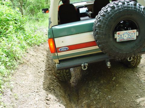 1996 Ford Bronco - 1996 Ford Bronco navigates a muddy off-road trail, spare tire visible on the back.
