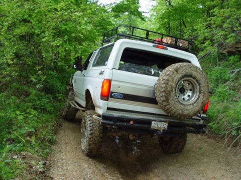 1996 Ford Bronco - A white 1996 Ford Bronco navigates a muddy, wooded trail with a spare tire on the rear and a loaded roof rack.
