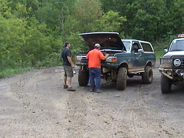 Green Ford Bronco with hood open, two men standing nearby. Another vehicle to the right