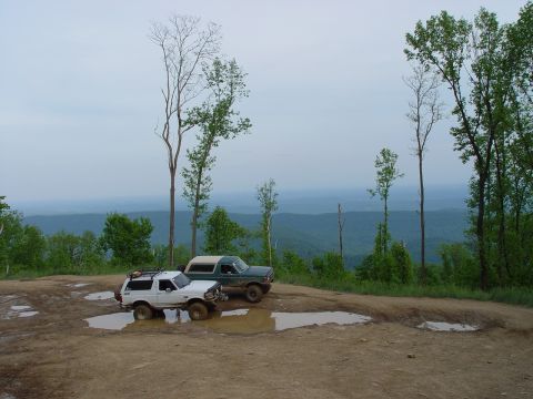 1996 Ford Bronco - Two SUVs navigate muddy terrain on a scenic overlook, with a vast expanse of green hills stretching into the distance.