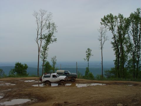 1996 Ford Bronco - Two SUVs, including a white 1996 Ford Bronco, are parked on a muddy, off-road hill with puddles and a scenic background.