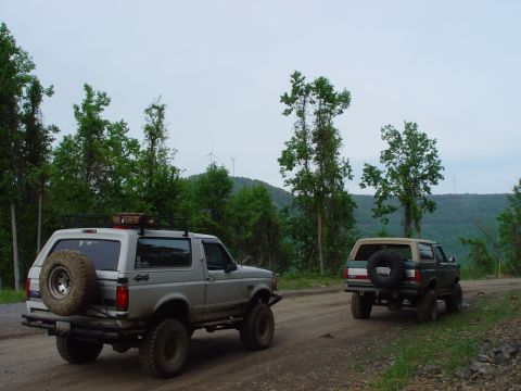 1996 Ford Bronco - Two Ford Broncos drive on a dirt road surrounded by trees and hills, with wind turbines visible in the distance.