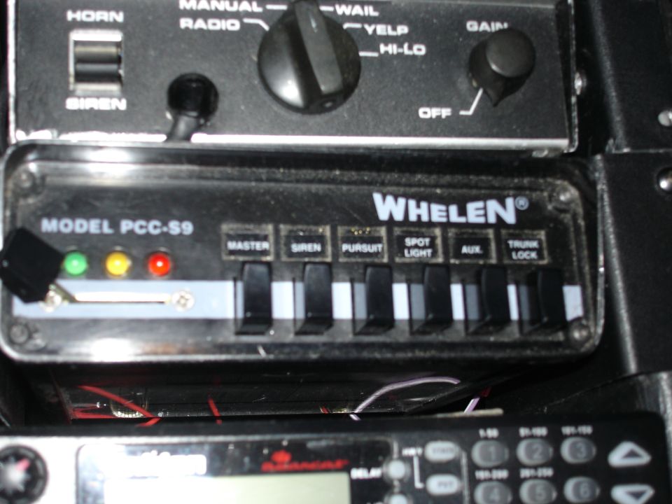 Close-up of a Whelen siren control panel with buttons for master, siren, pursuit, spot light, aux, and trunk lock. Also shows a horn and radio control