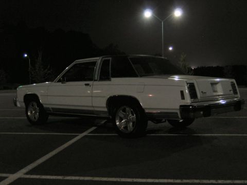 1985 Ford LTD Crown Victoria - Night shot of a white two-door Ford full-size coupe photographed from the driver's-side rear three-quarter in an empty parking lot under so…