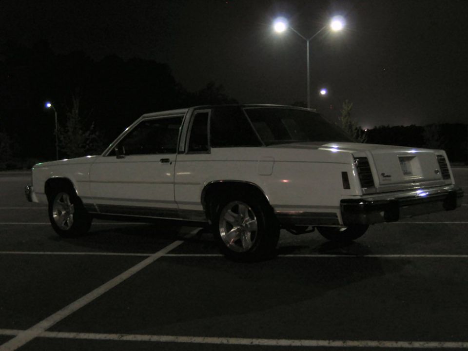 White two-door Ford coupe at night in parking lot, rear three-quarter view on five-spoke alloys