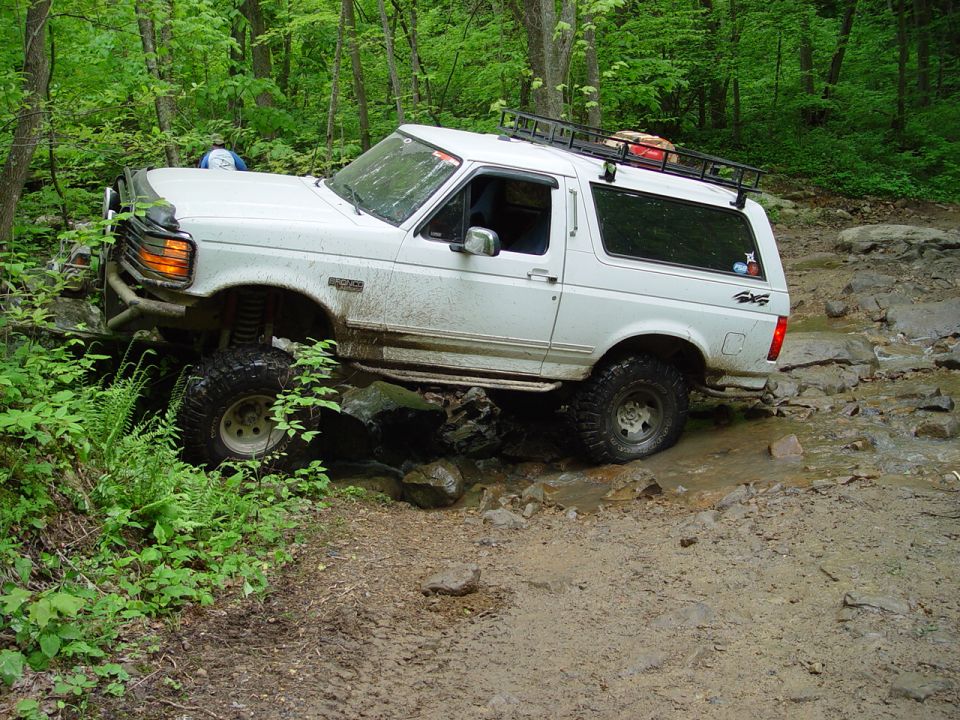 1996 Ford Bronco navigating a rocky, muddy off-road trail with trees on either side