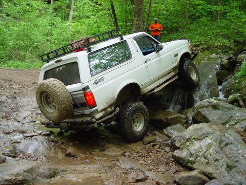 1996 Ford Bronco - A white 1996 Ford Bronco tackles a challenging off-road trail, driving through a rocky stream.