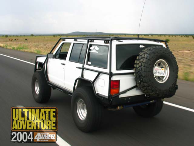 Lifted white Jeep Cherokee XJ with exterior cage and rear tire carrier driving on a highway