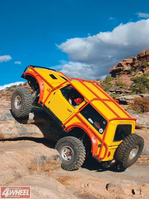 1996 Ford Bronco - A heavily modified yellow off-road vehicle navigates a rocky terrain under a blue sky with scattered clouds.